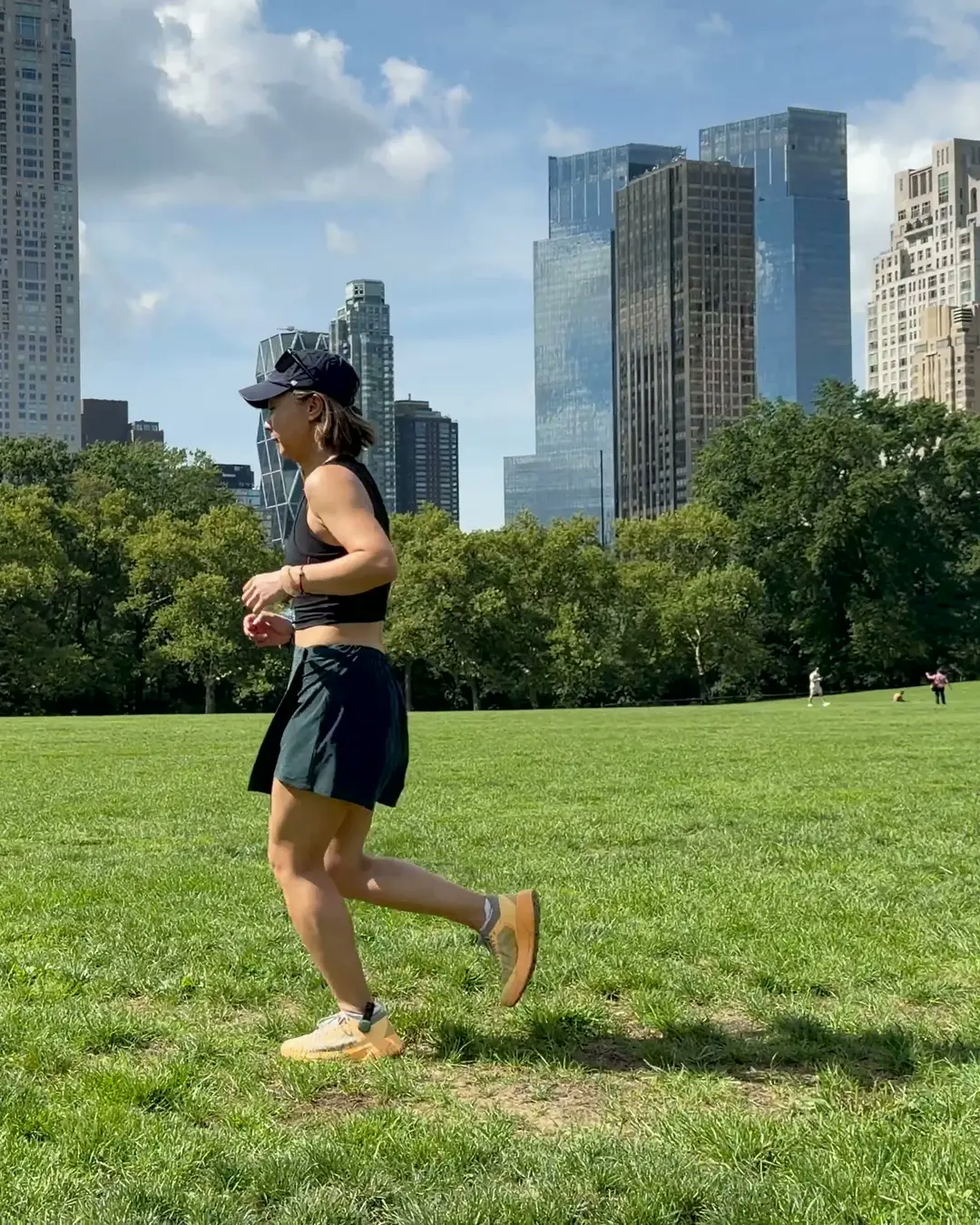 A runner mid-stride across the grass in Central Park, with the Midtown skyline behind her.