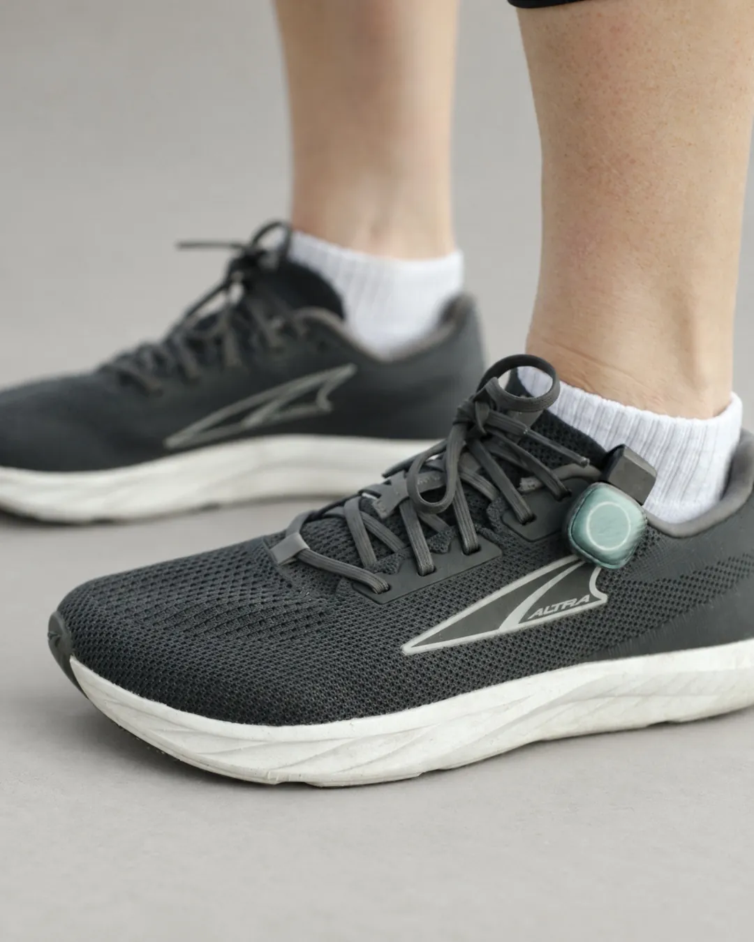 A runner's black running shoes on a grey floor, a small STRIV pod clipped to the laces.